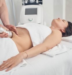 Calm smiling young lady lying on the medical couch with towels on her and a cosmetologist applying vacuum suction to her belly