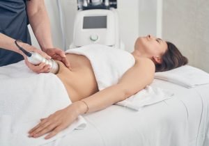 Calm smiling young lady lying on the medical couch with towels on her and a cosmetologist applying vacuum suction to her belly